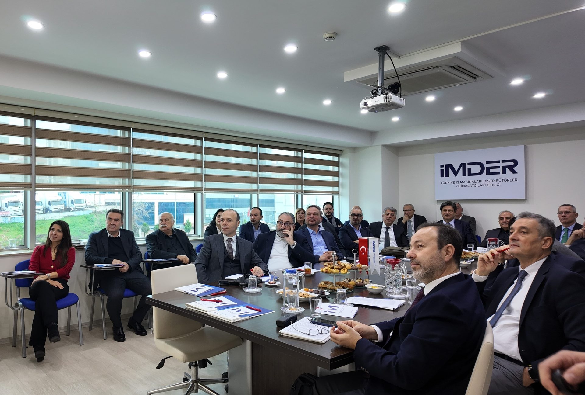 A group of business professionals gathered around a conference table for a meeting, with a projector and screen.