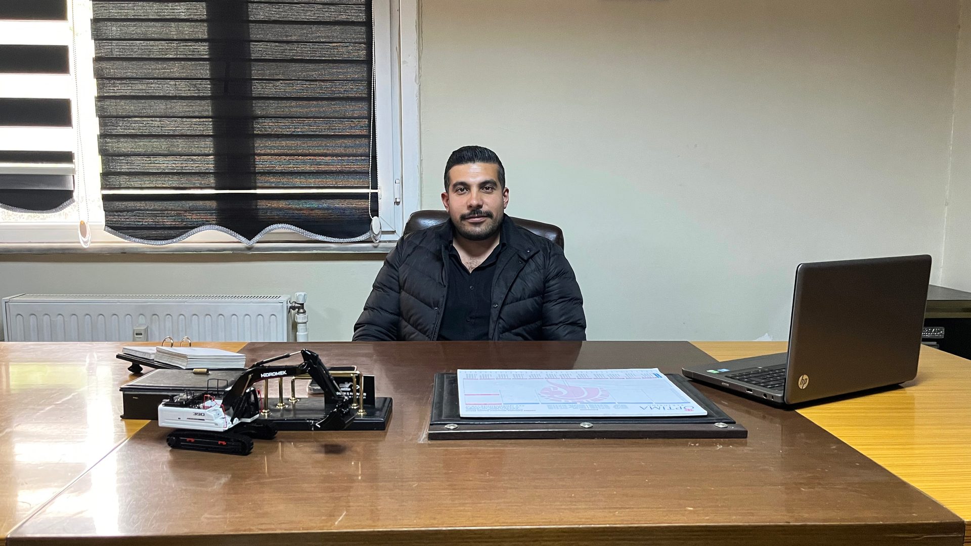 Man in black jacket sits at office desk with laptop and miniature excavator.