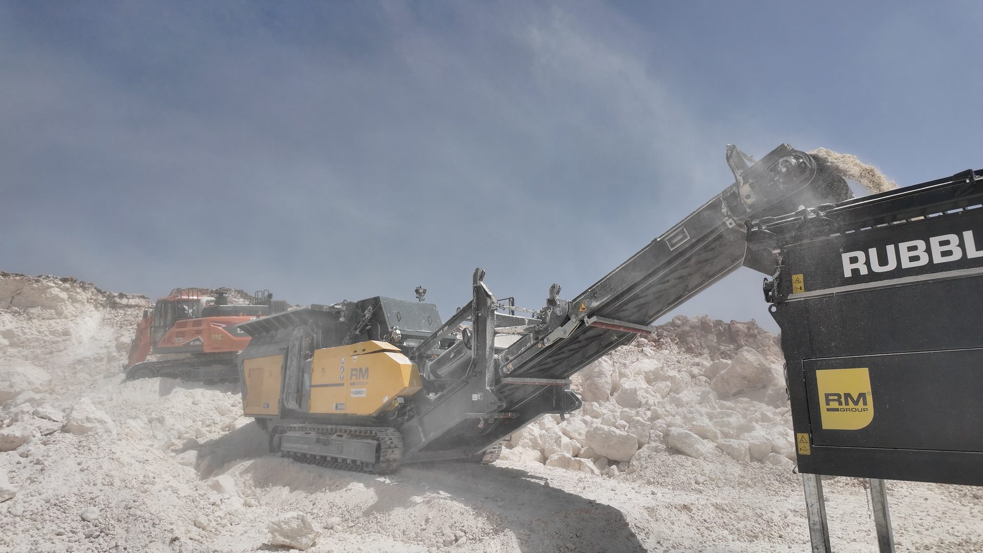RM crusher processing white rock in dusty quarry, excavator in background.