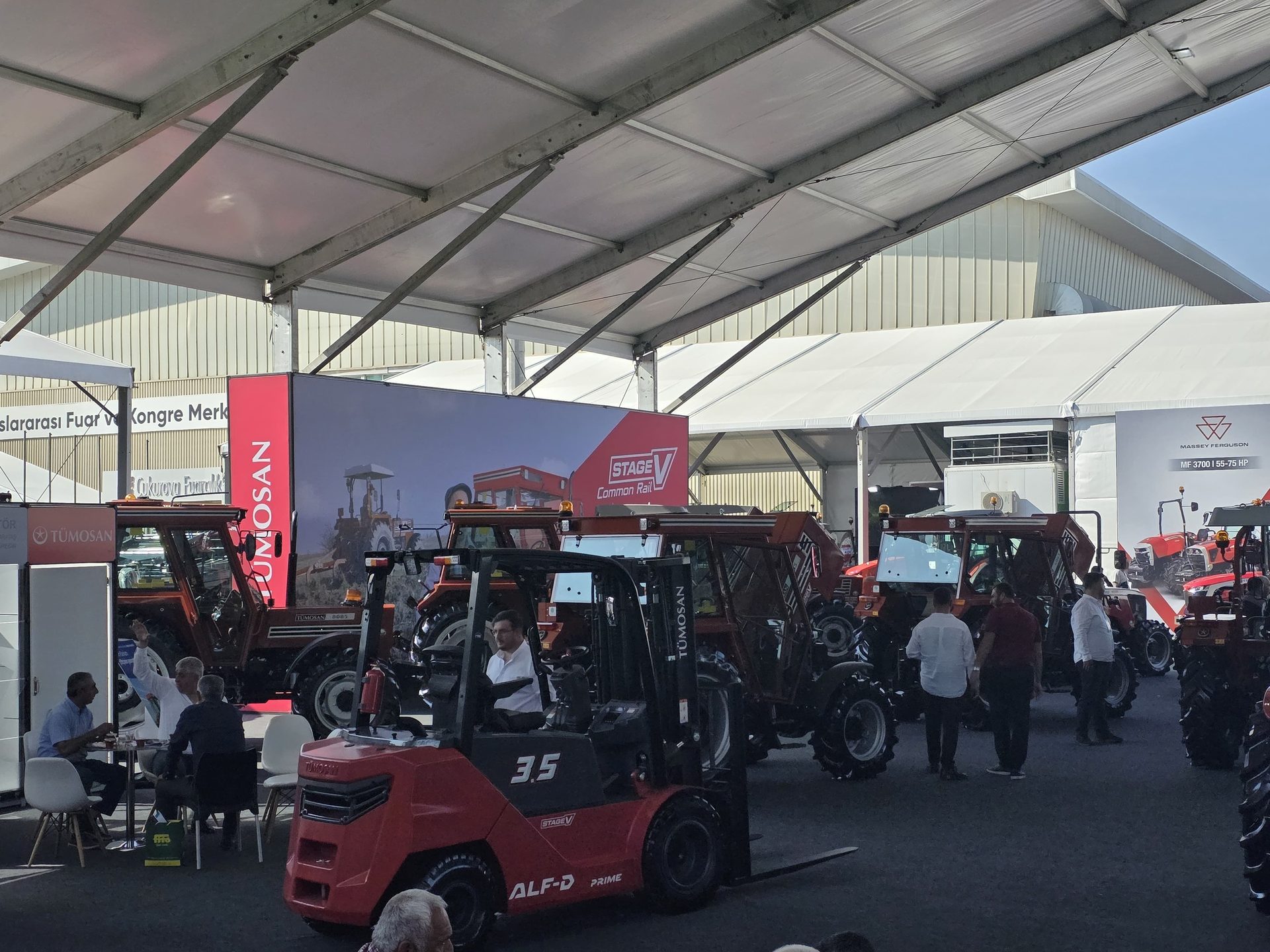 Trade show with tractors, a red forklift, and people under large white tents.