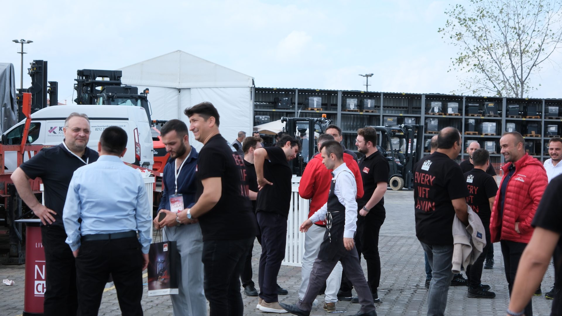 Men gathered outdoors at an industrial event with forklifts and a white tent in the background.