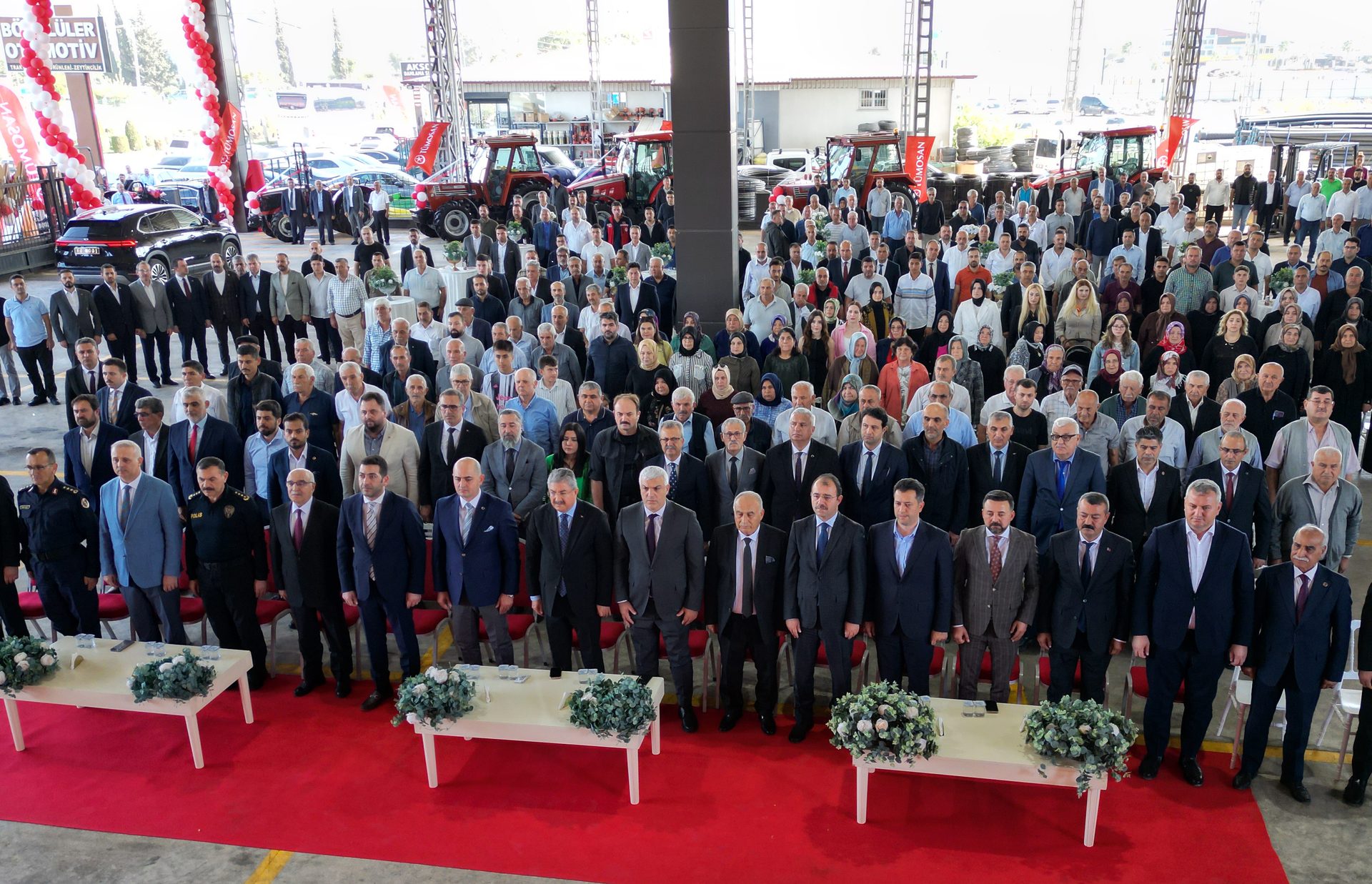 Formal gathering of people, mostly men, inside a showroom with tractors and cars.