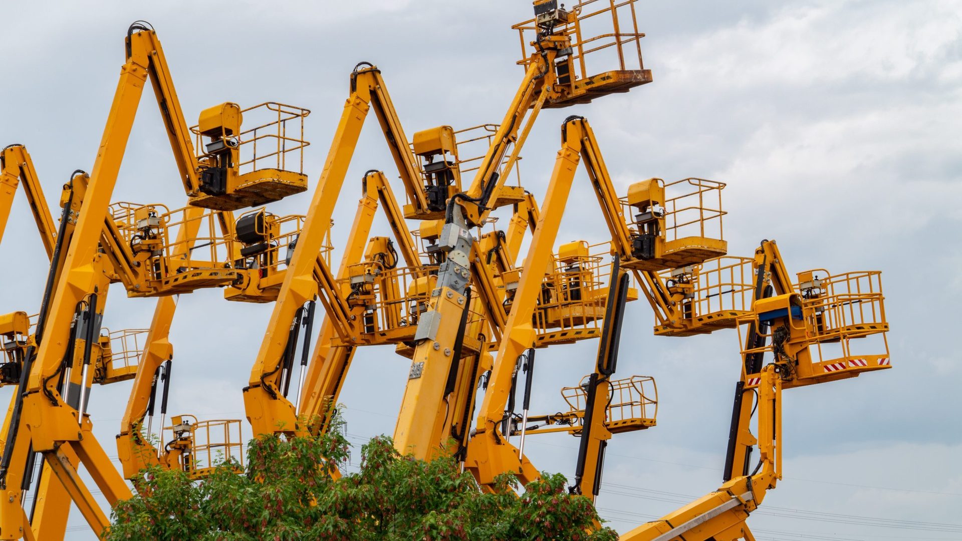 Many yellow cherry pickers against cloudy sky, trees below.