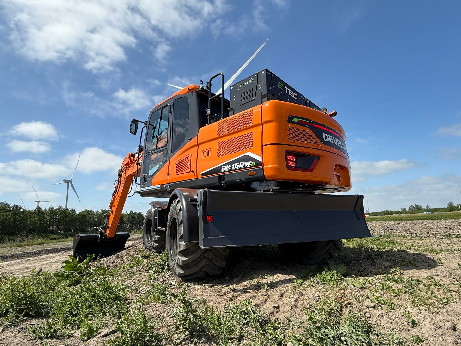 Orange electric excavator on a job site with wind turbines in the background.