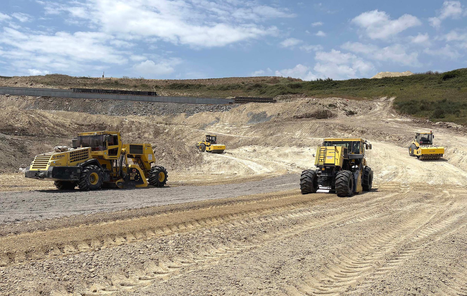 Yellow Bomag heavy machinery on a dirt road construction site under a cloudy sky.