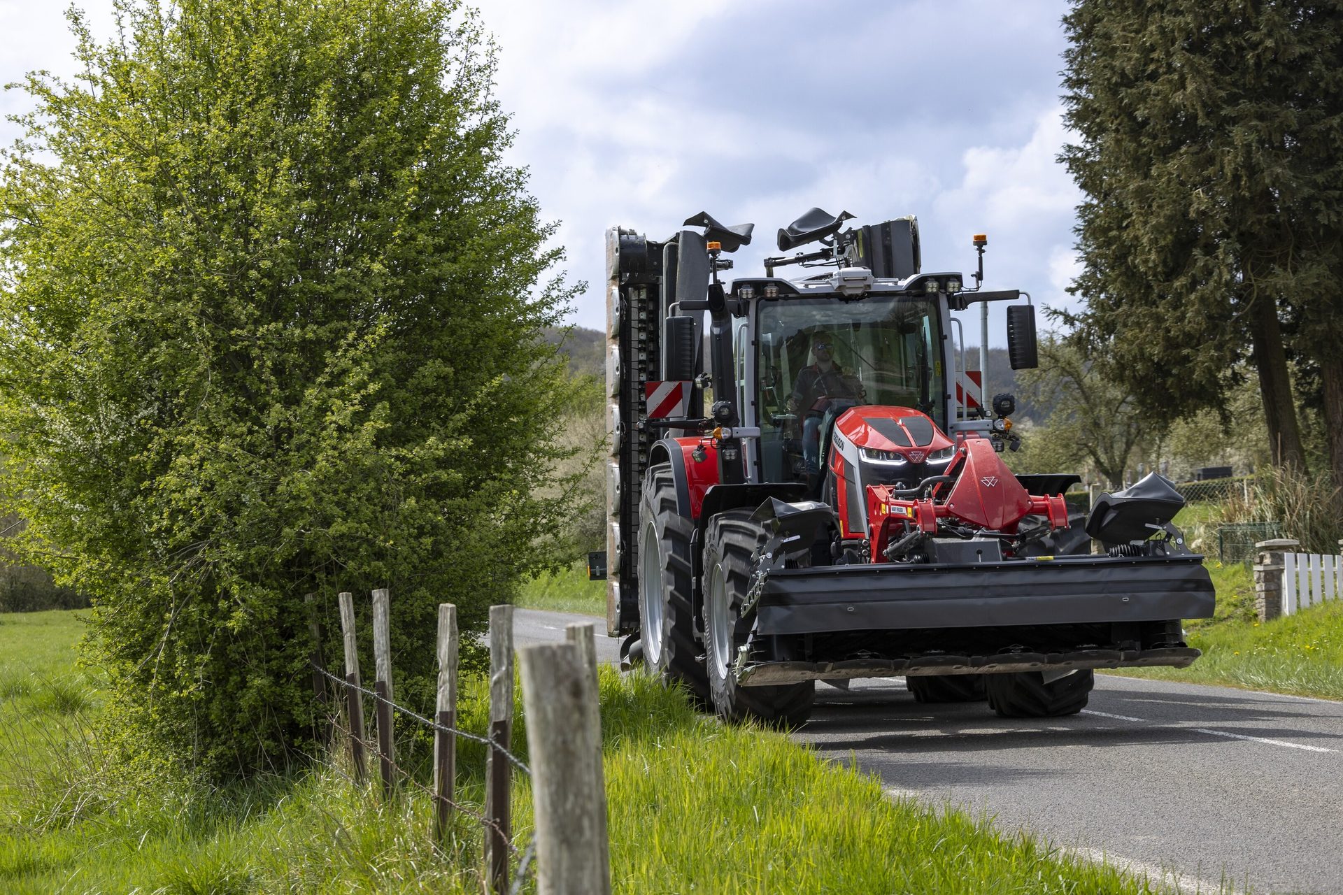 Red and gray tractor with large implement drives on a road next to a green tree.
