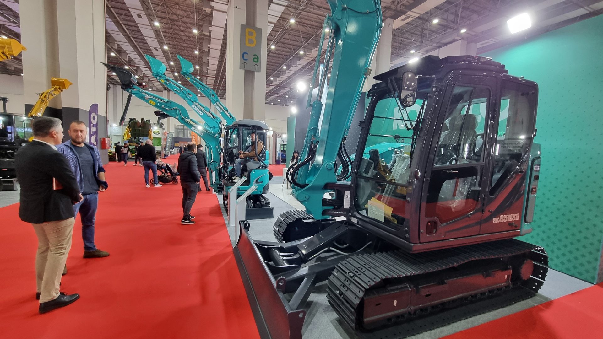 Teal excavators on display at an exhibition, with people walking on a red carpet.