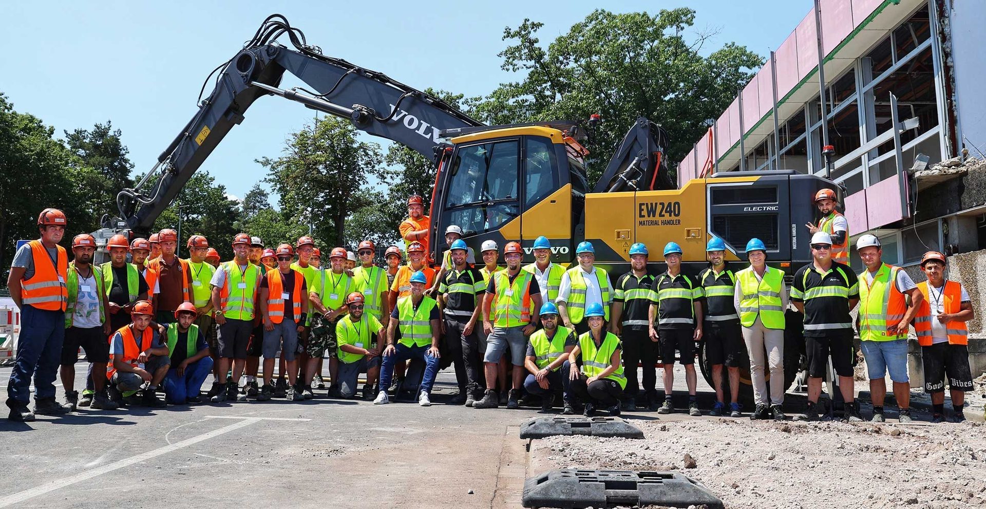 Construction workers and staff pose with a large Volvo electric excavator at a demolition site.
