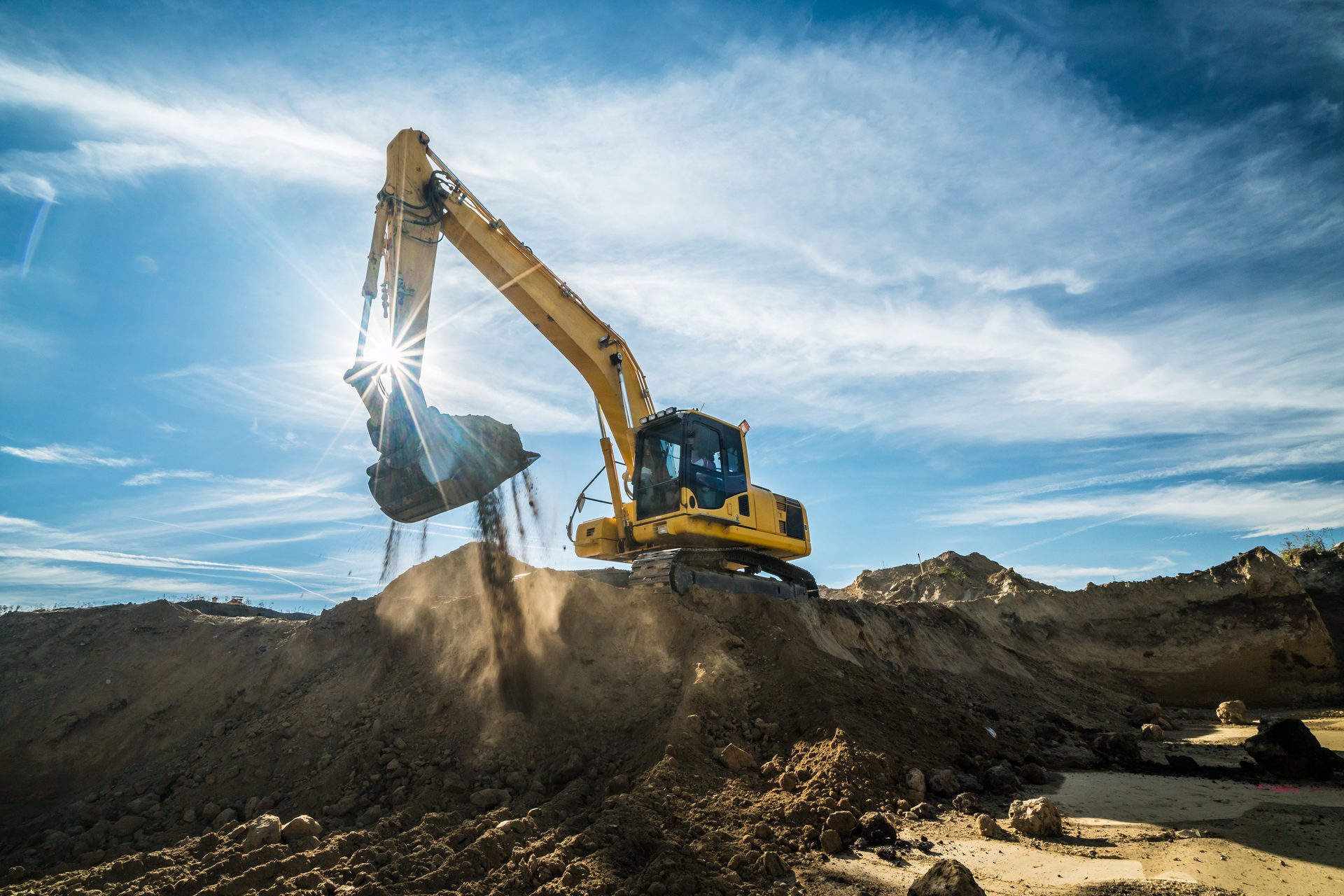Yellow excavator dumping soil, bright sun with lens flare over a dusty construction site.