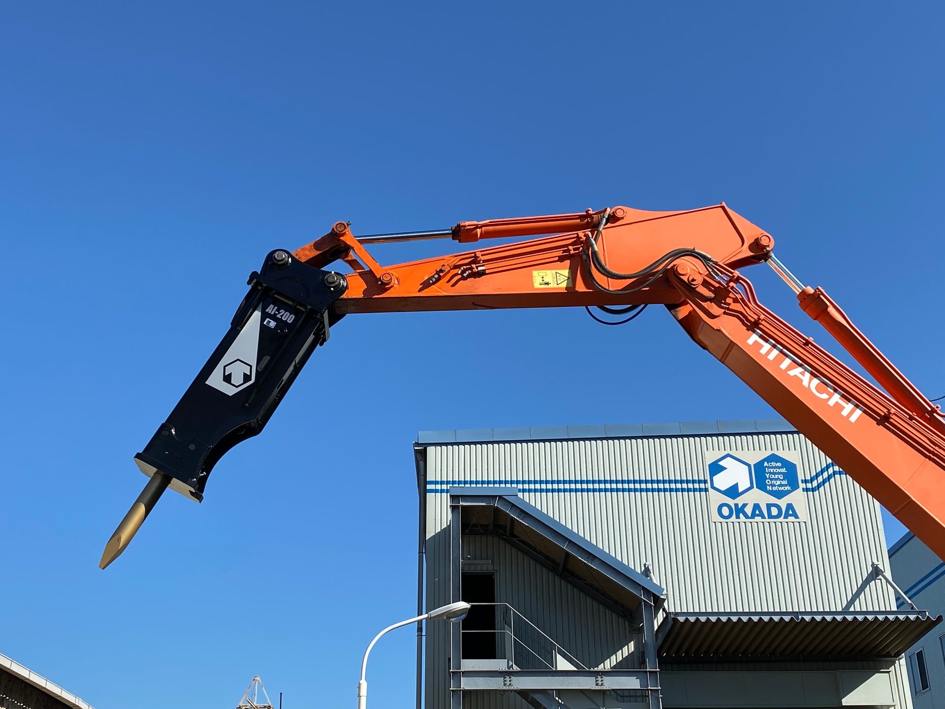 Hitachi excavator with Okada AF-200 hydraulic breaker against blue sky and Okada building.
