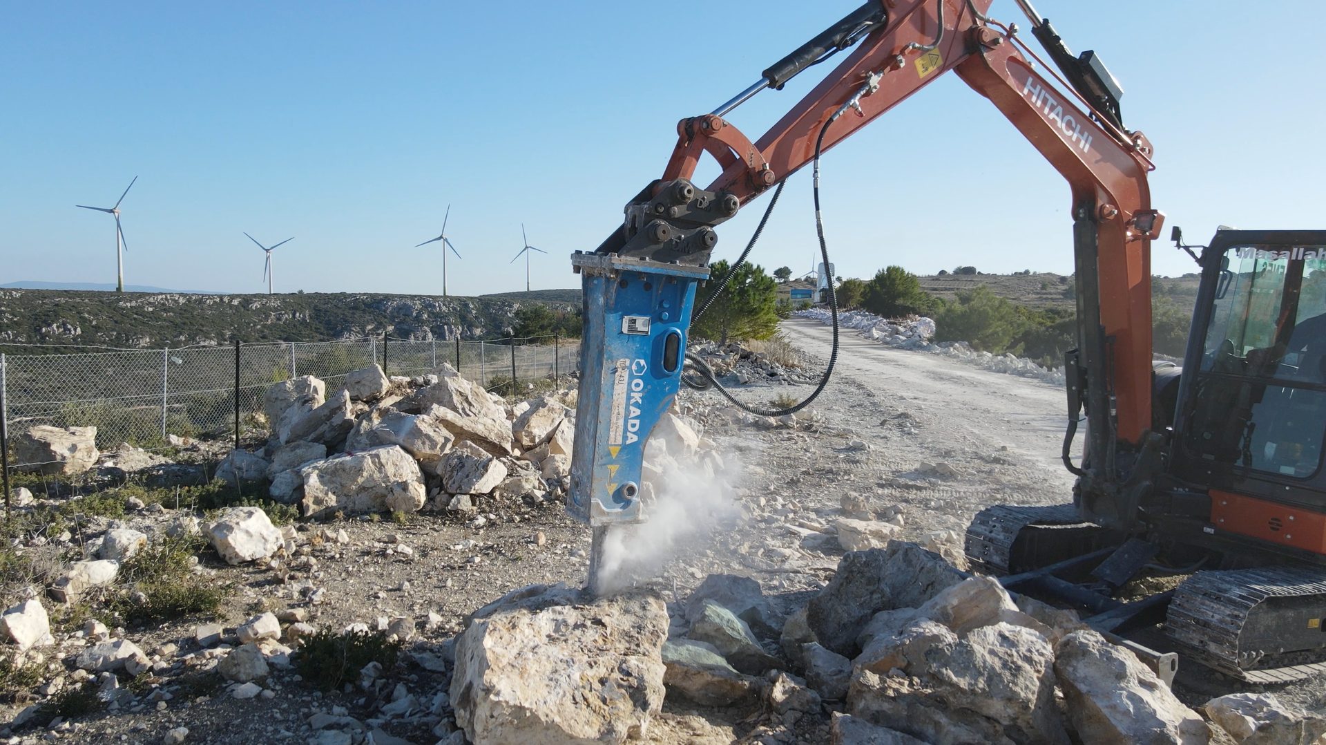A Hitachi excavator with an Okada hydraulic breaker demolishes rocks on a construction site near wind turbines.