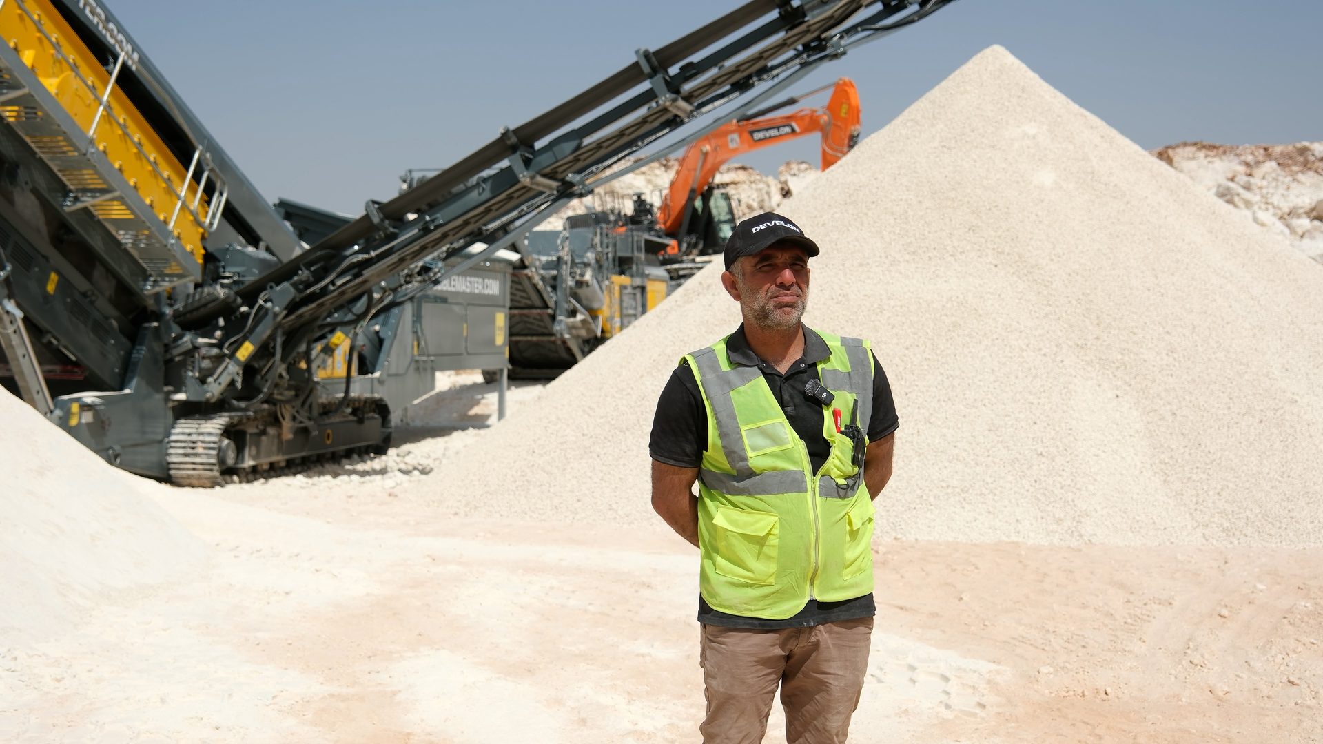 Man in high-vis vest at a quarry with rock crushing machinery and aggregate piles.