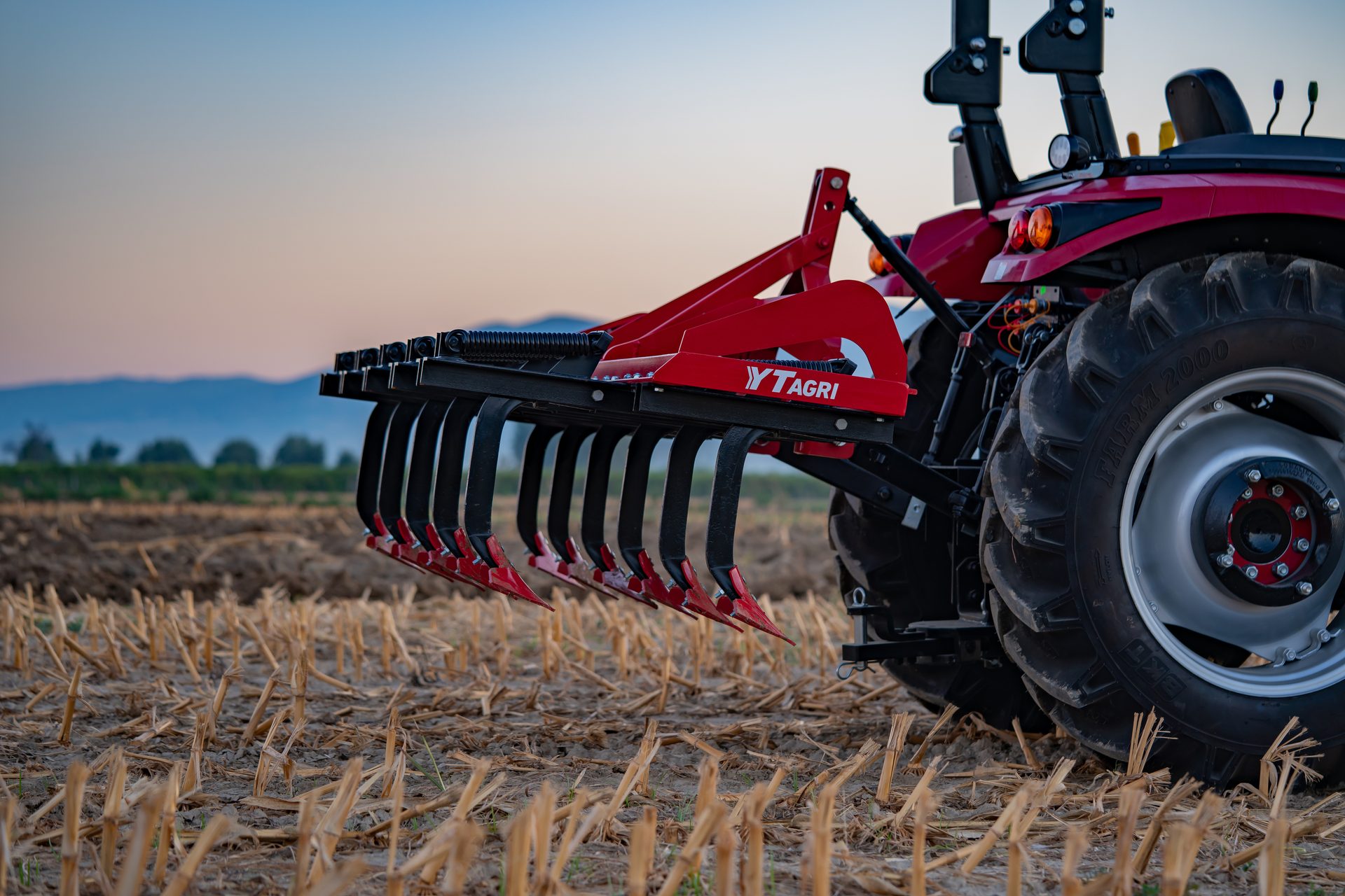 Red YTAGRI agricultural cultivator attached to a tractor, set in a field with harvested stalks under a clear sky.