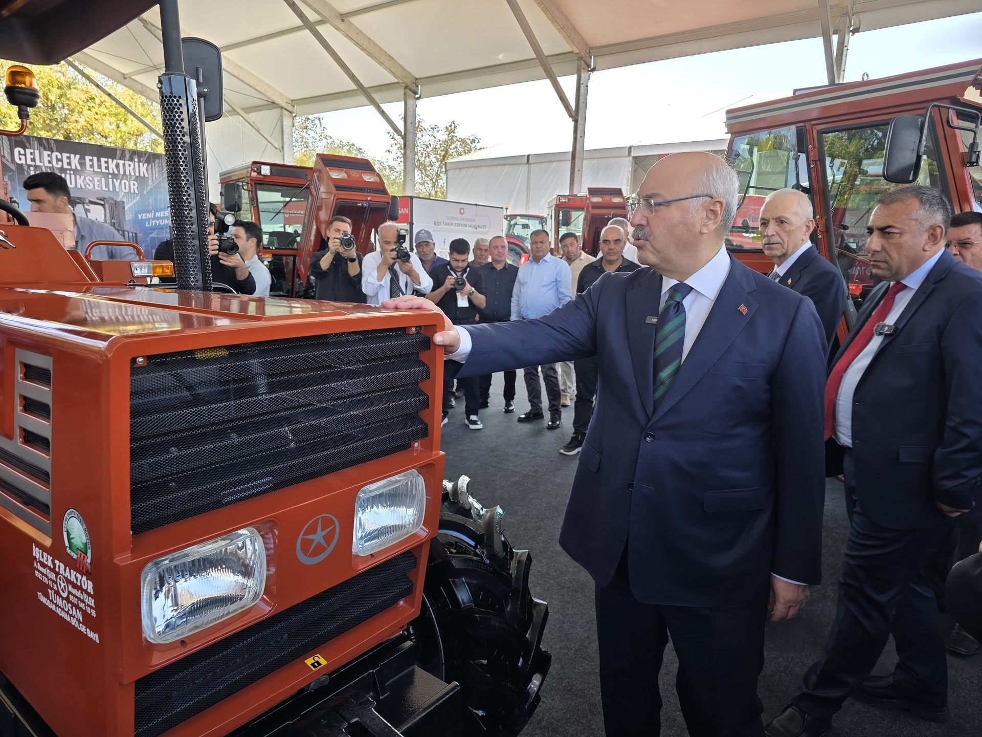 A man in a suit touches an orange tractor at an exhibition, with photographers in the background.