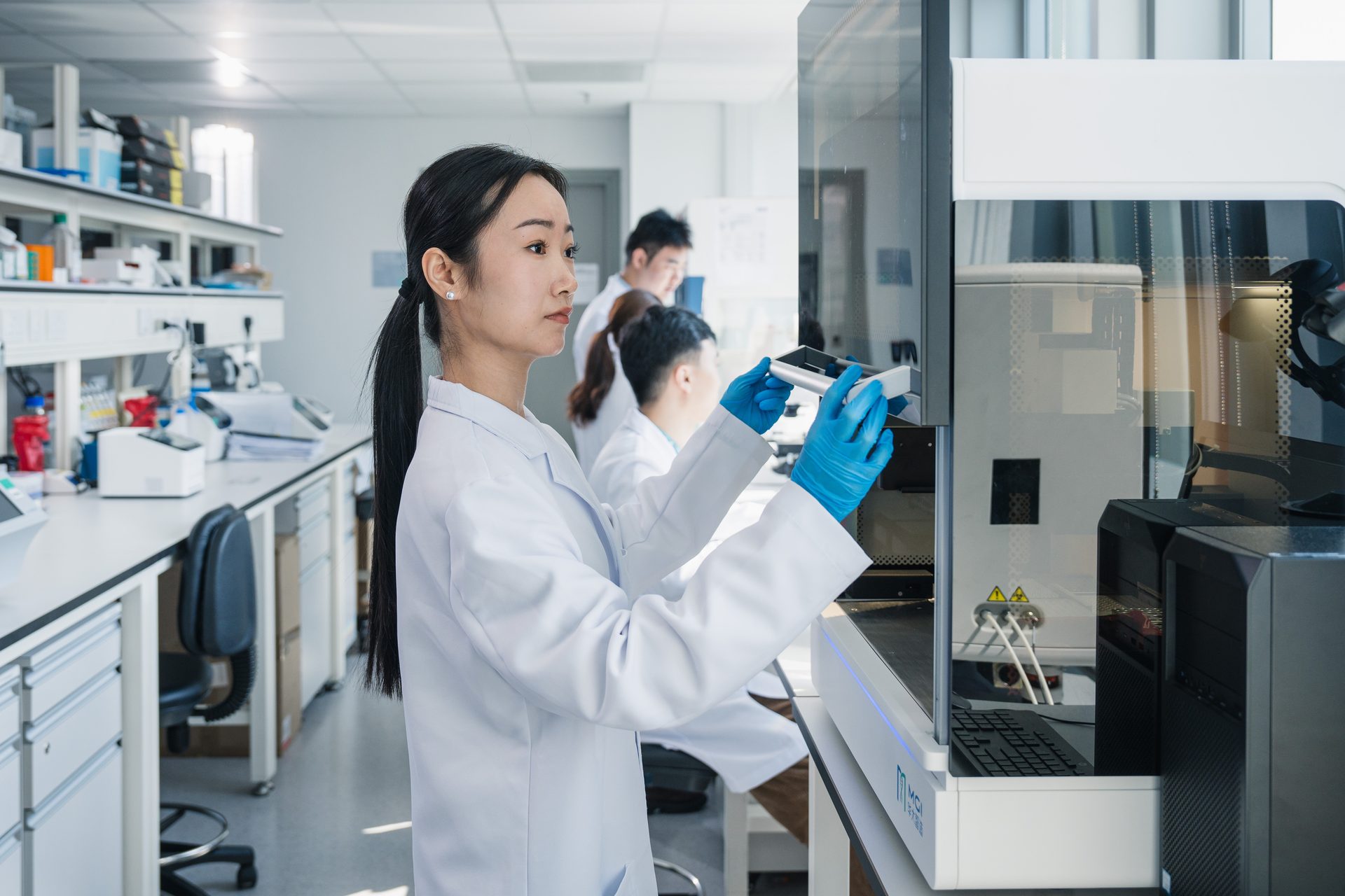 Asian woman scientist in lab coat and gloves operating high-tech lab equipment, with other researchers nearby.
