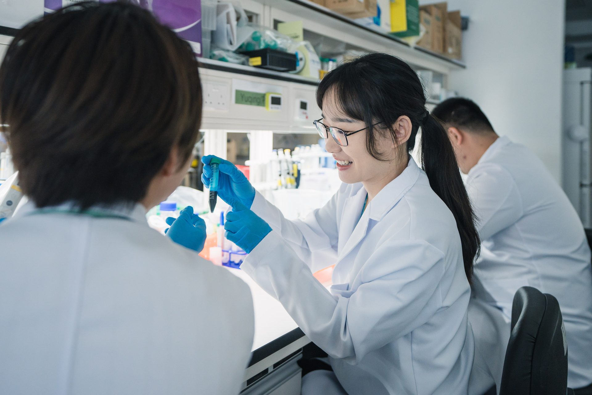Two scientists in lab coats and gloves collaborating, examining a test tube with blue liquid.