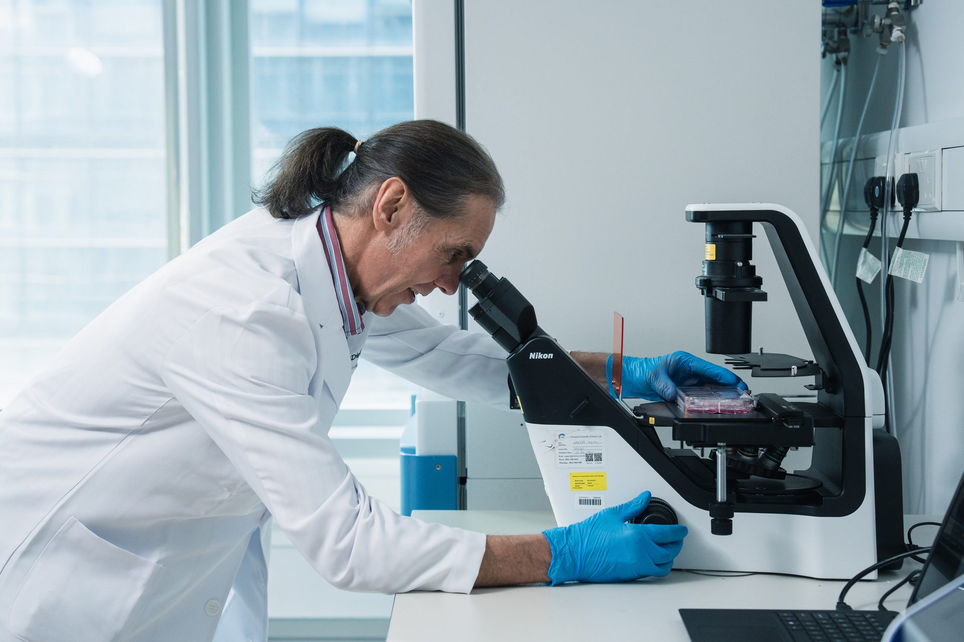 Scientist in a lab coat and blue gloves examining samples under a Nikon microscope.