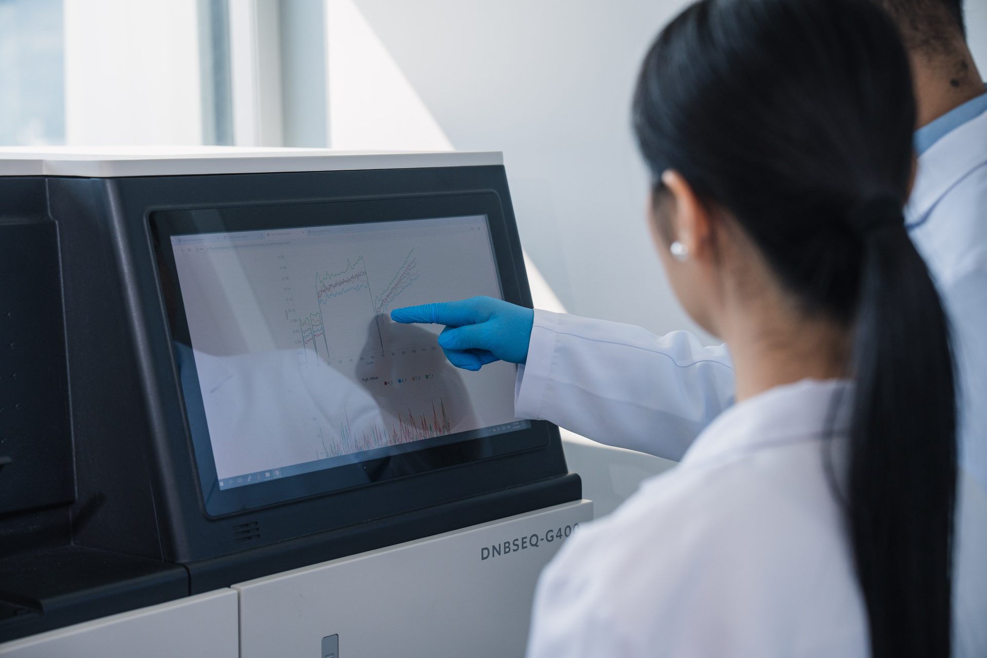 A scientist in a lab coat and blue glove points at graphs on a large screen of a DNA sequencer.
