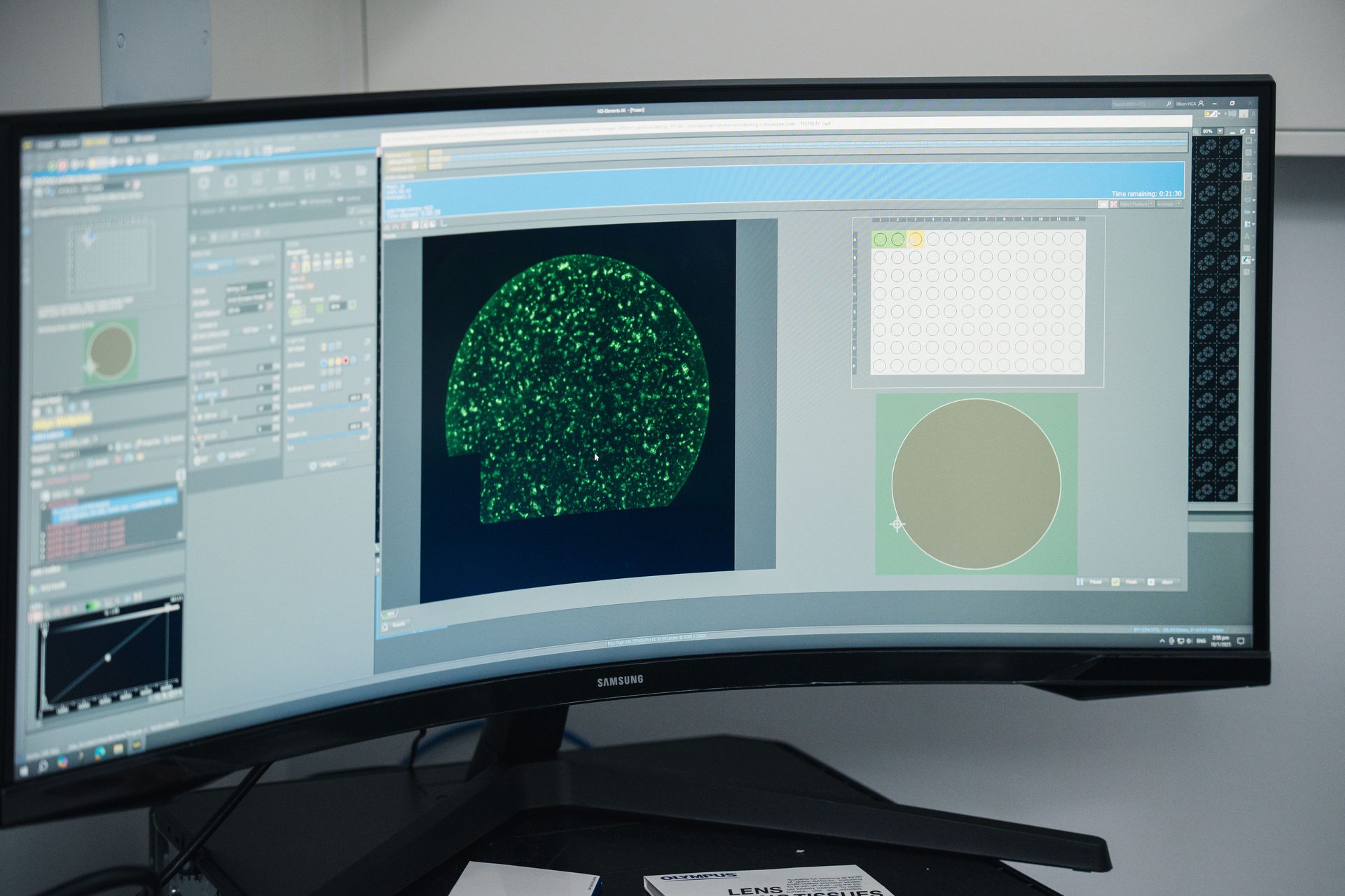 A curved monitor displays scientific software with a green microscopic image of cells and various control panels.