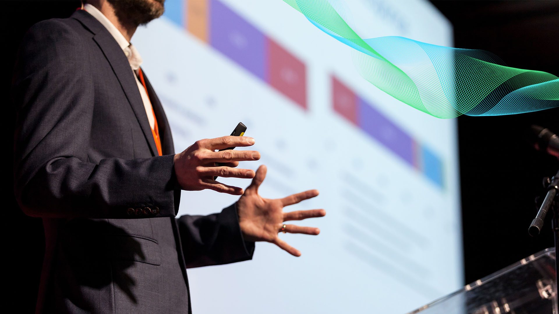 Presenter in suit gesturing, screen with data graphics and wavy lines.
