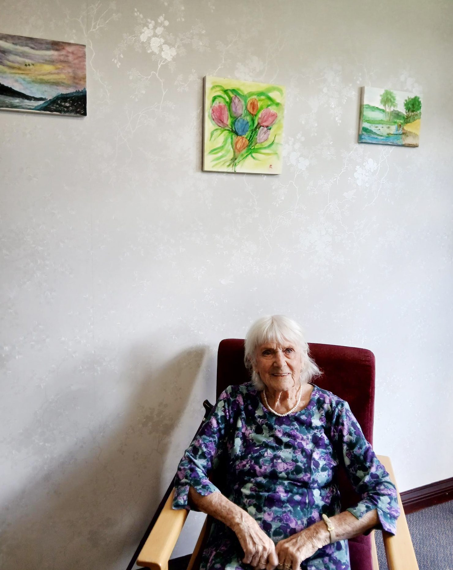 Smiling resident Edith seated in a chair, with three artworks on a patterned wall.