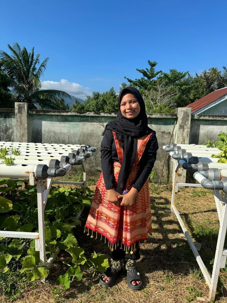 A young woman in a black hijab and traditional attire smiles in a sunny outdoor hydroponic garden.