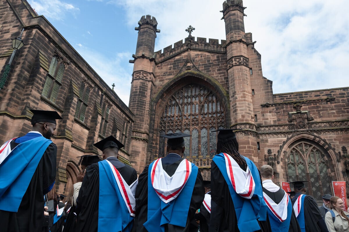 Graduates in academic robes approach a grand, gothic stone building with intricate windows.