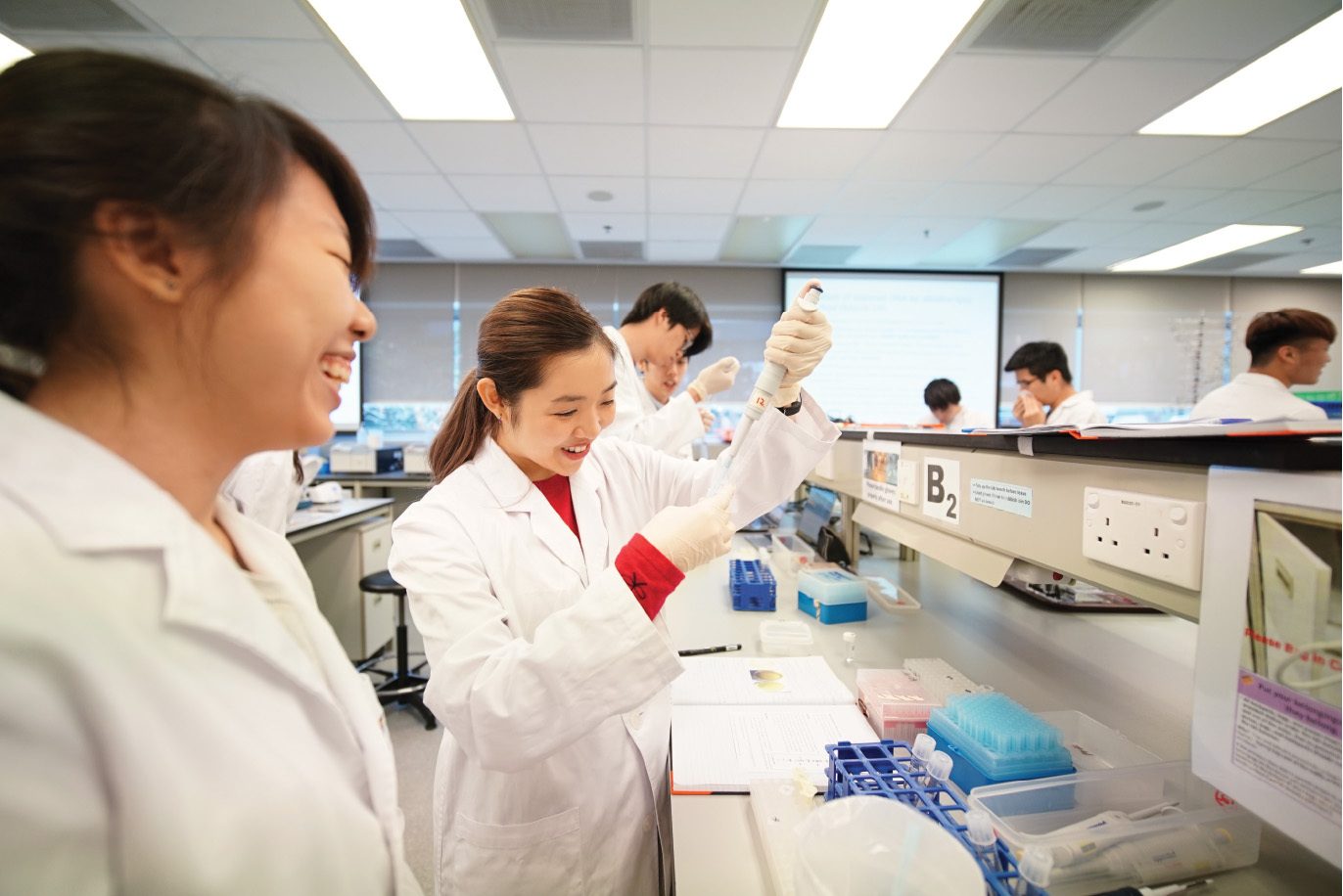 Two smiling female students in a lab, one pipetting.