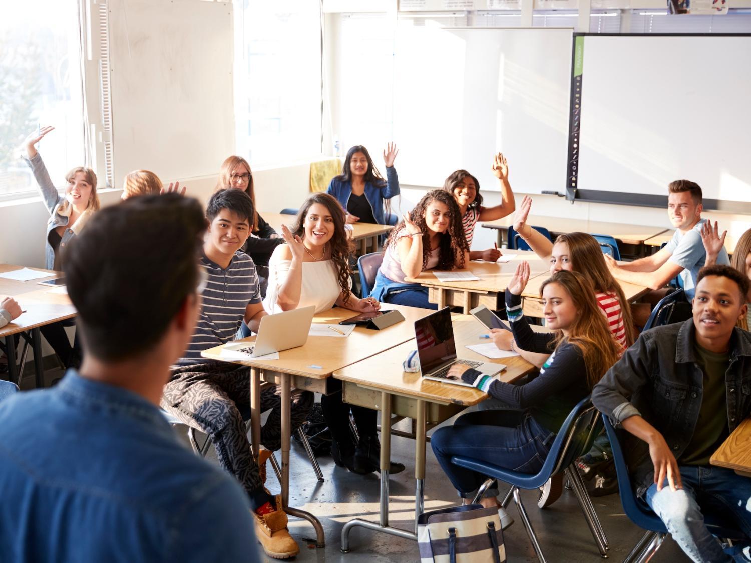 Diverse students in a classroom raising hands, facing a teacher in the foreground.