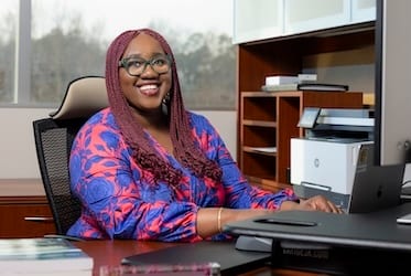 Smiling Black woman with braids and glasses at an office desk, wearing a blue and red floral top.