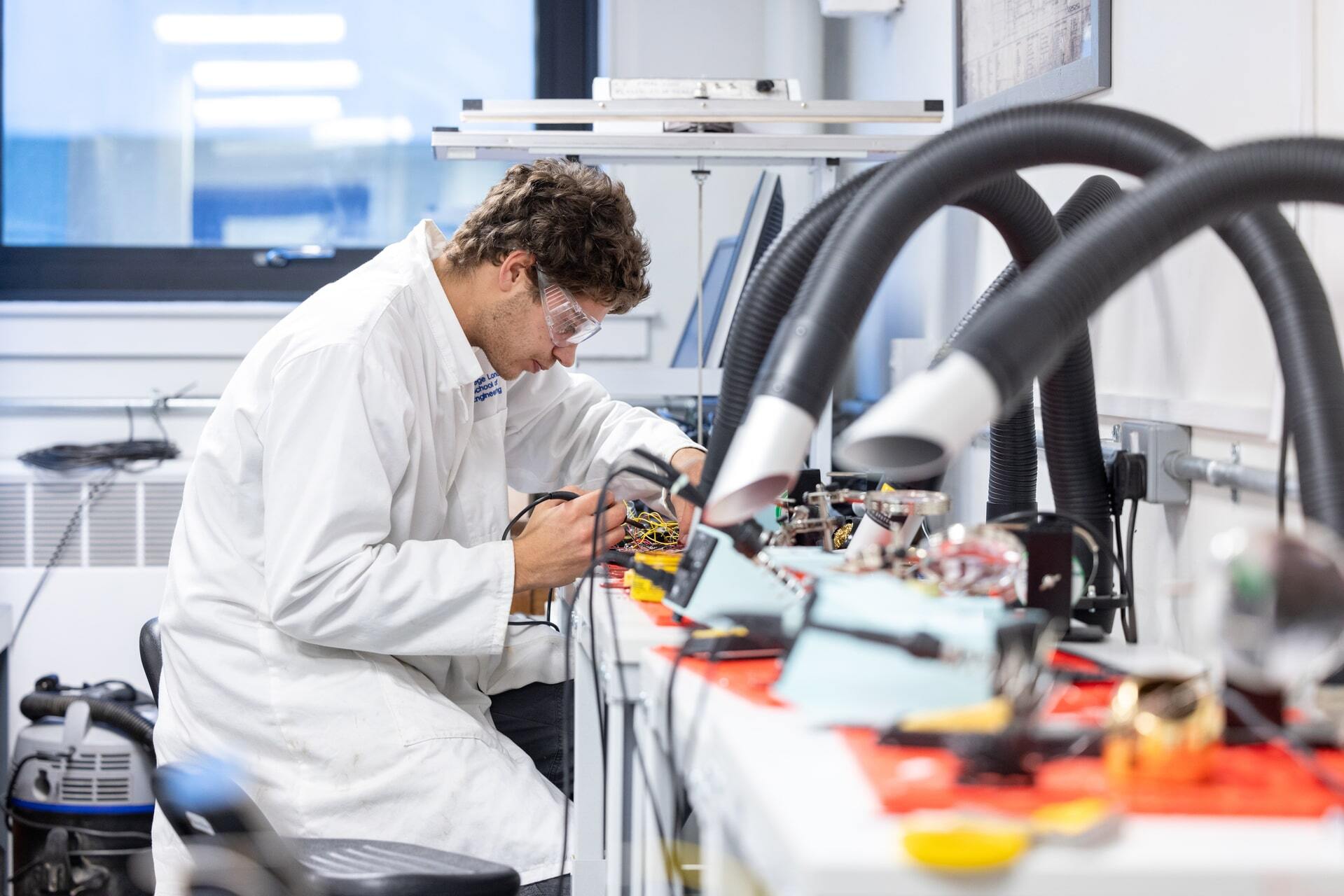 A man in a lab coat and safety glasses works on electronics in a lab.