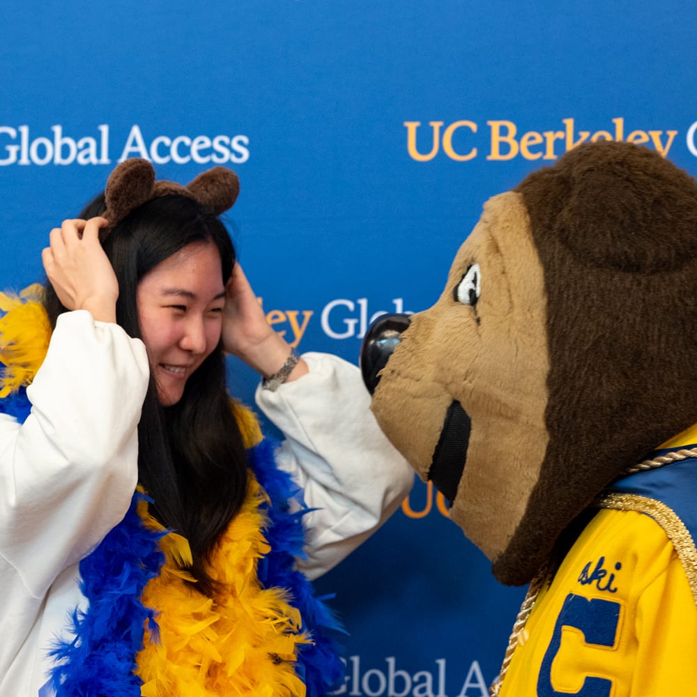 Smiling woman with animal ears and blue/yellow boa next to UC Berkeley mascot.
