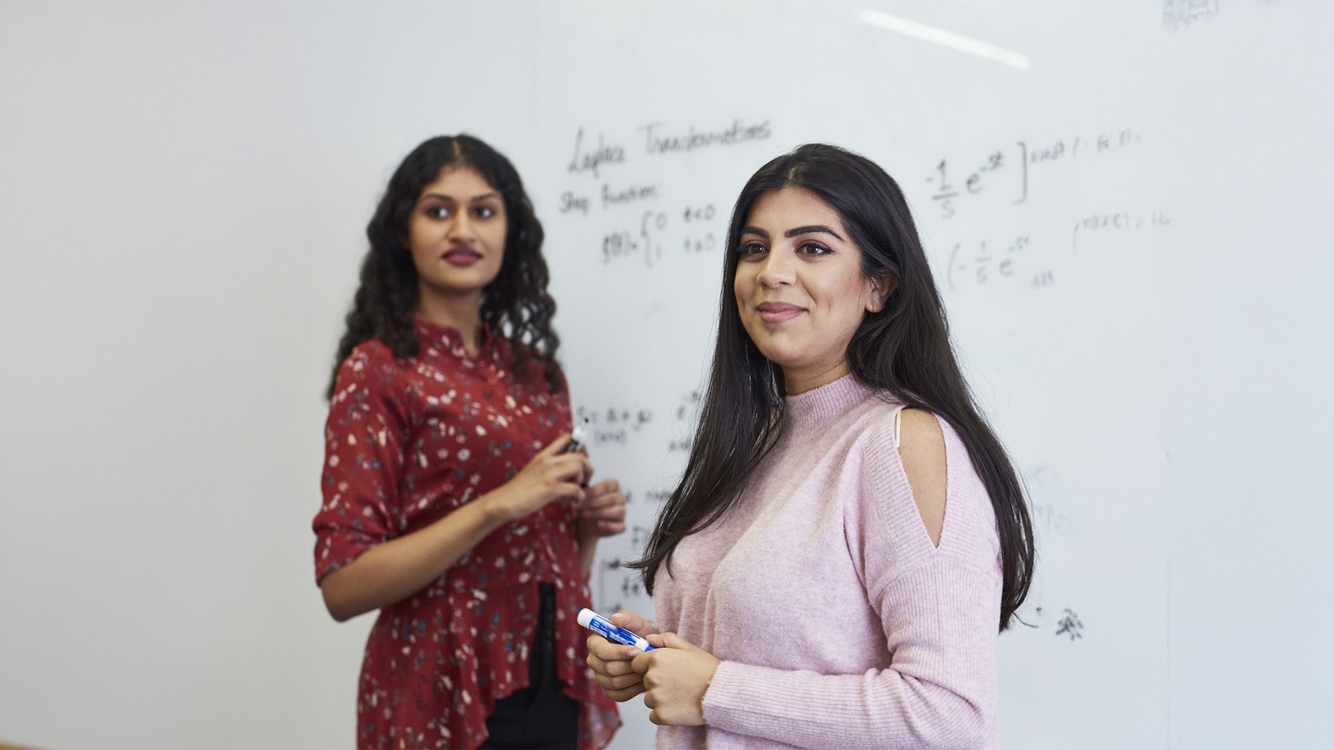 Two women stand by a whiteboard with math equations, one smiling and holding a marker.