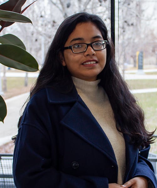 Smiling young woman with glasses, long dark hair, wearing a dark blue coat and a light turtleneck.
