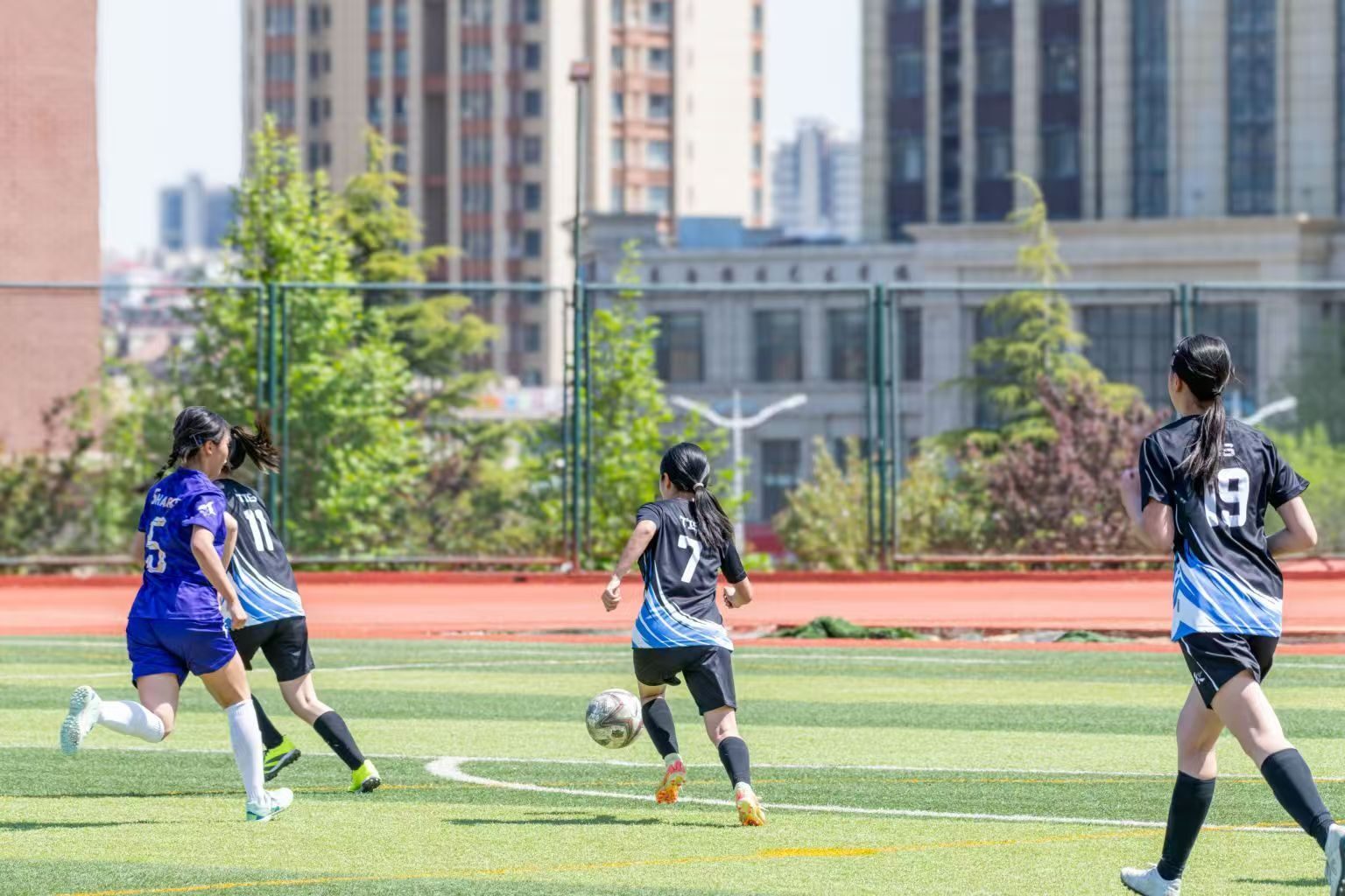 Women play soccer on a green field; a player dribbles, with an urban background.