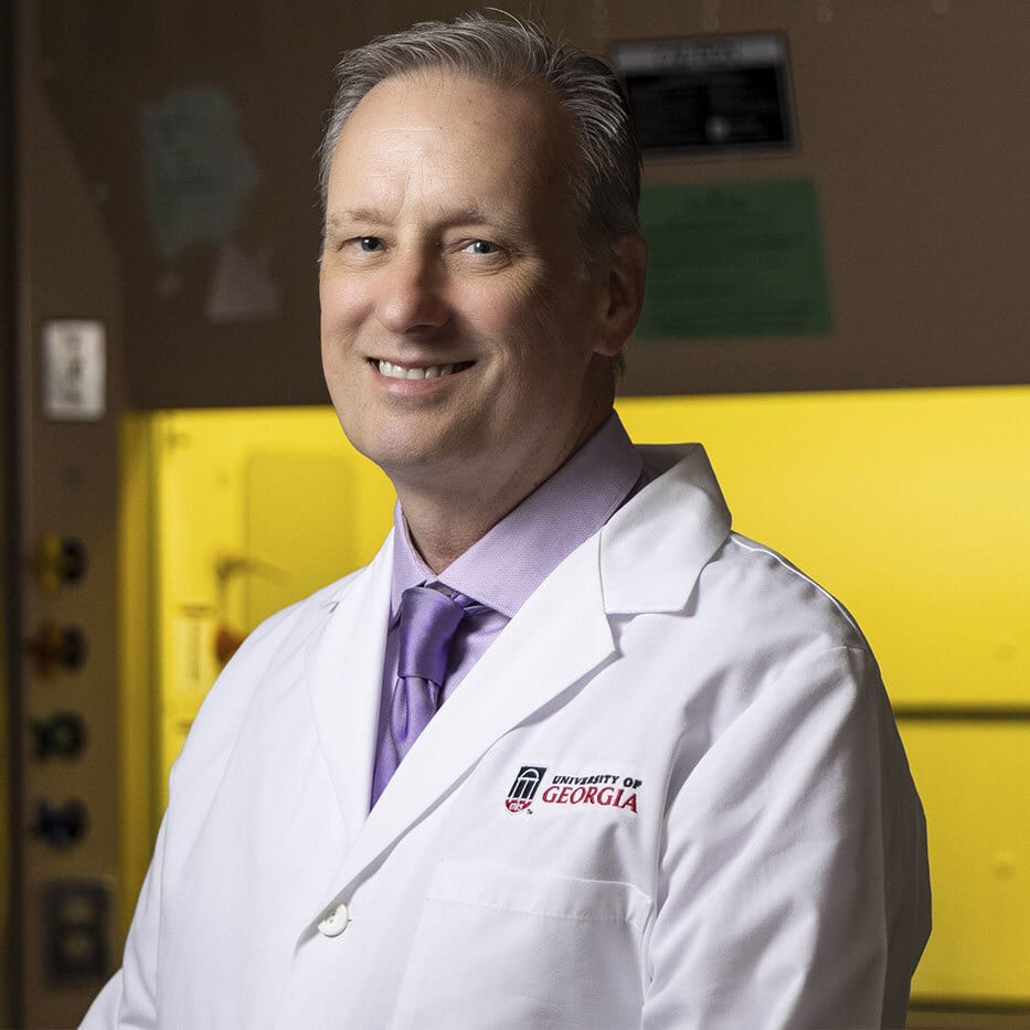 A smiling man in a lab coat and purple tie in a lab setting.