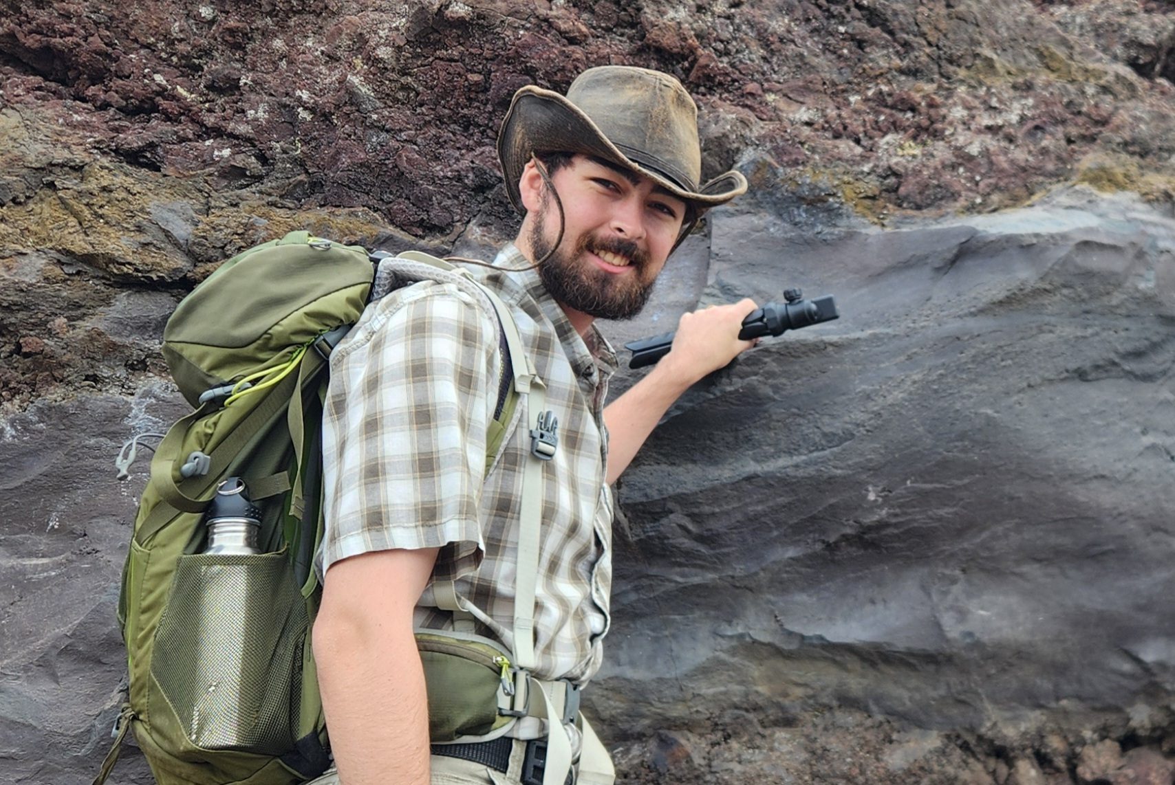 Smiling bearded man in hat and backpack, holding rock wall.