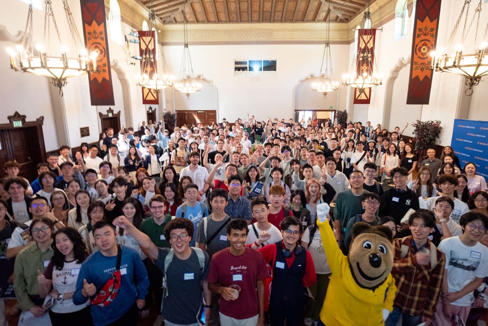 Many happy students and a bear mascot filling a large, ornate hall, smiling and waving.