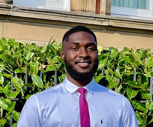 A smiling young Black man in a white shirt and pink tie, standing outdoors in front of a hedge.