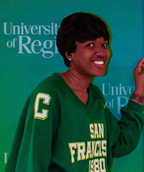 Smiling Black woman in green "San Francisco 1980" sweatshirt, "University of Regina" backdrop.