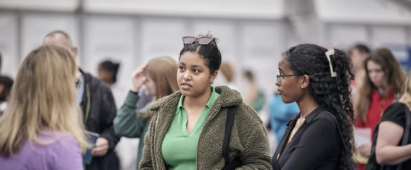 Two young women, one with sunglasses on her head, stand in a diverse indoor crowd.