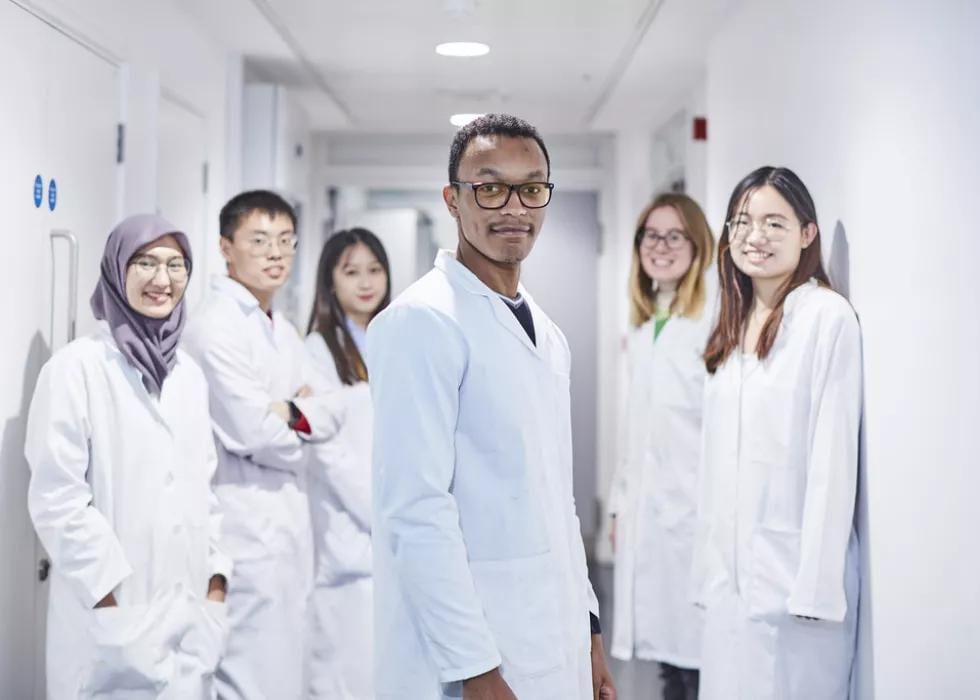 Diverse team of six scientists in lab coats, smiling in a hallway.
