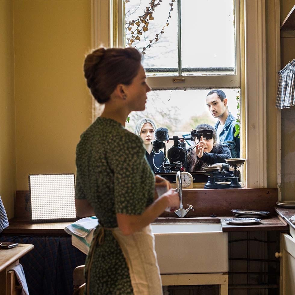 A woman in a period kitchen, being filmed by a crew visible through the window.