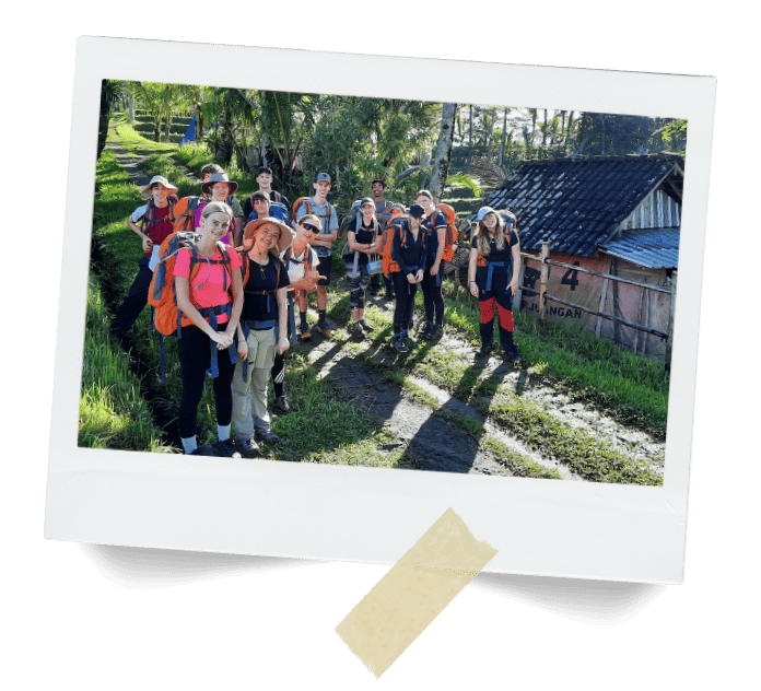 A group of young hikers with backpacks on a dirt path, with tropical trees and a small building.