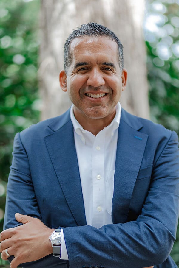 A smiling man in a blue suit and white shirt with his arms crossed, standing outdoors.