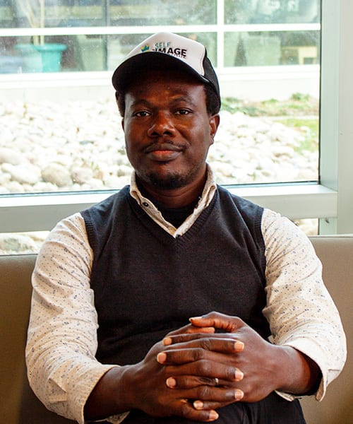 A man in a "SELF IMAGE" cap and vest, hands clasped, looking at the camera.