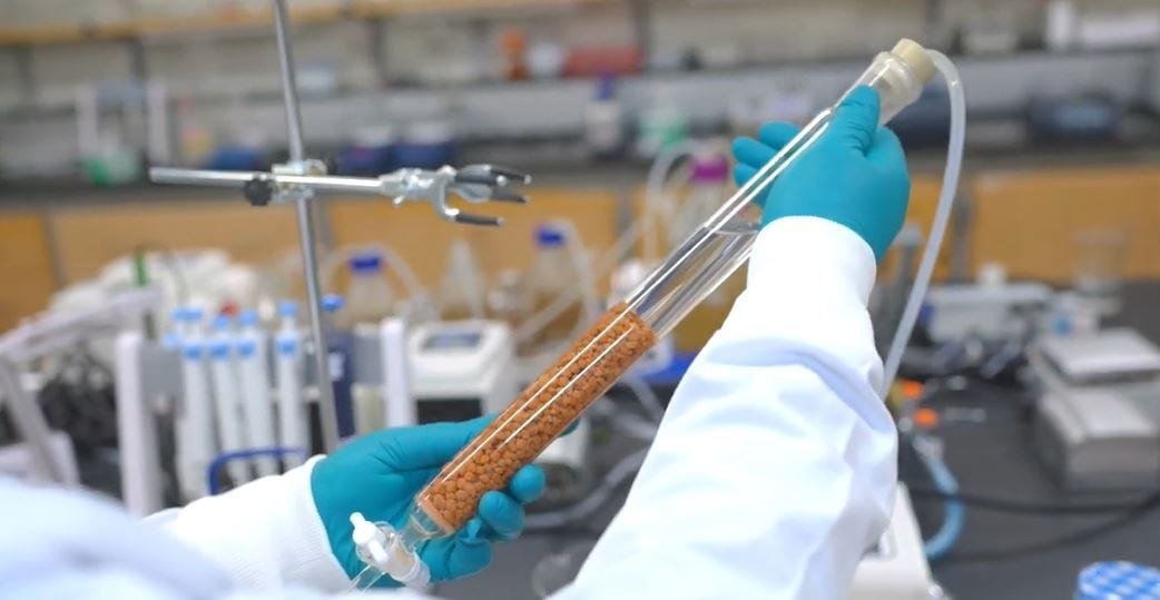 Scientist in lab gloves holds a clear column packed with orange-brown beads.