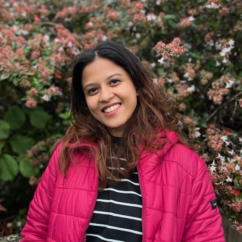 A smiling woman in a pink puffer jacket and striped shirt, against a blurred background of foliage and flowers.