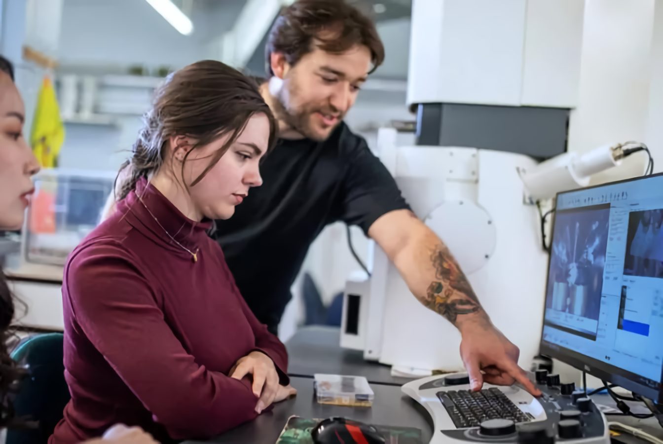Three lab scientists examining data on a computer screen, one pointing.