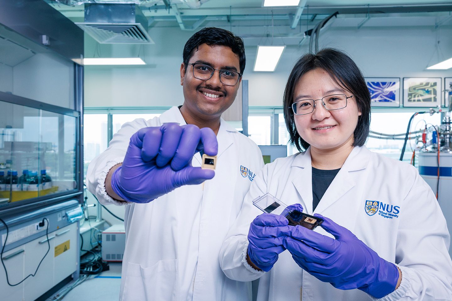 Two scientists in lab coats and gloves smiling, holding small electronic components in a laboratory.